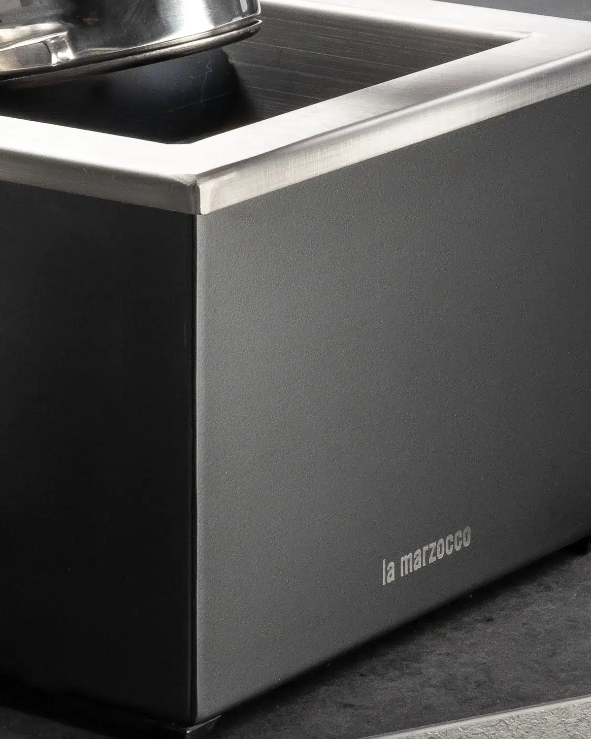 Corner of the black La Marzocco knock box with its brushed steel rim and etched wordmark