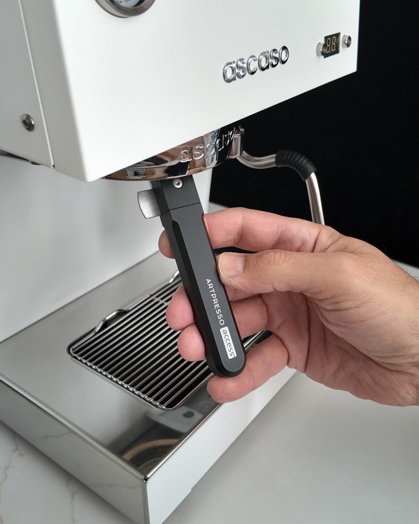 Hand gripping the Artpresso Access tool locked into the group head of a white Ascaso espresso machine, above the chrome drip tray