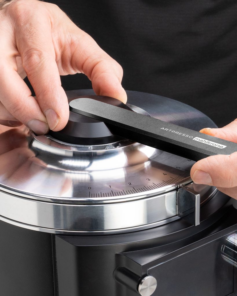 Hands laying the Artpresso Mahlkrone wrench across the burr collar of a black Mahlkonig grinder, the adjustment scale visible below