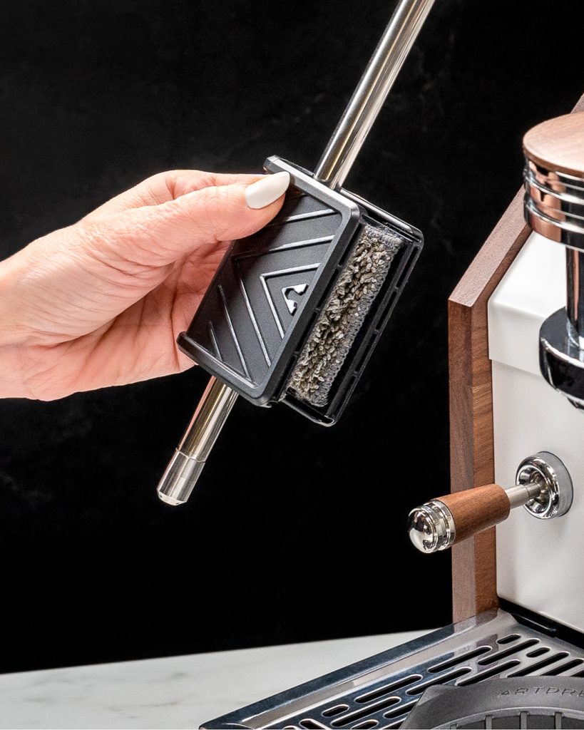 Hand holding the steam wand cleaning tool closed around the steam wand of a white espresso machine with walnut side panels