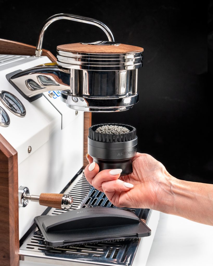 Hand raising the 58mm group head cleaning tool, brush upward, toward the group of a white espresso machine with walnut side panels