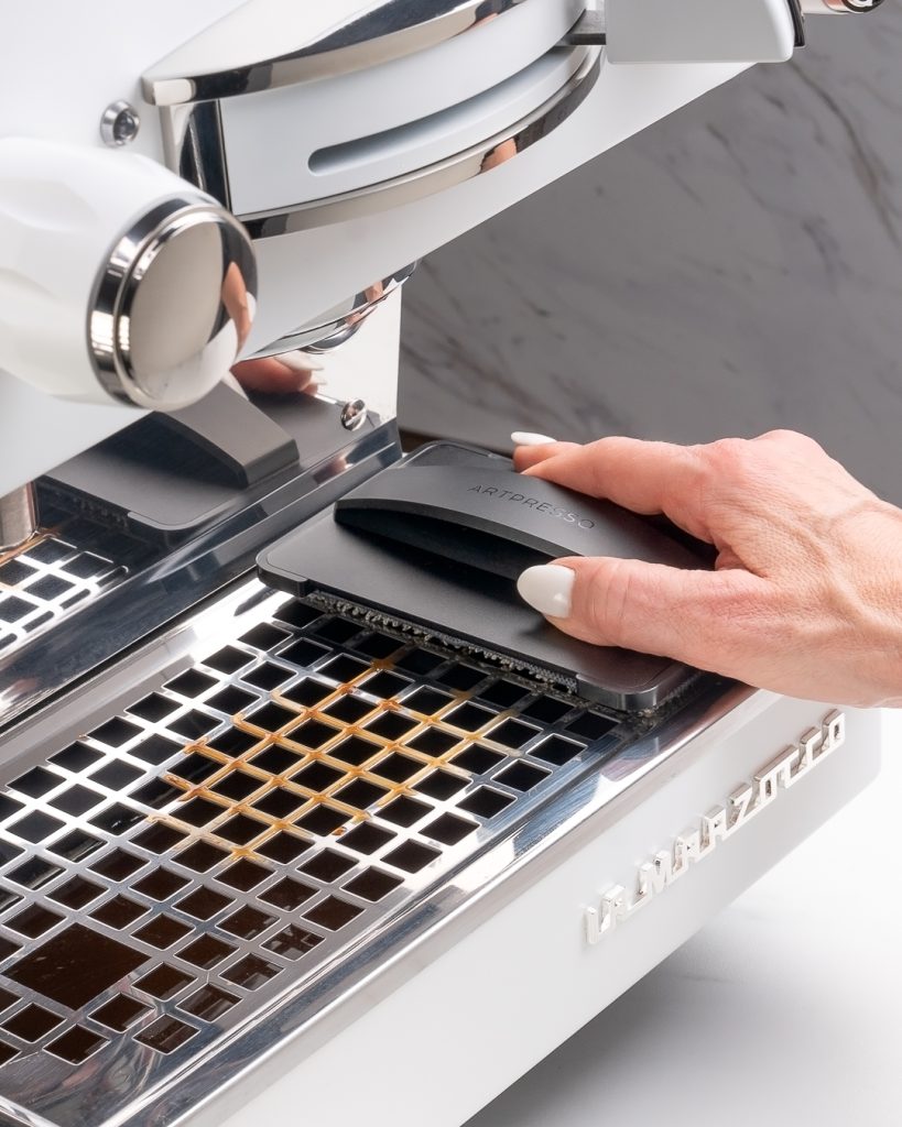 Hand drawing the Drain Tray Pro cleaning tool across the chrome drip tray grate of a white La Marzocco espresso machine