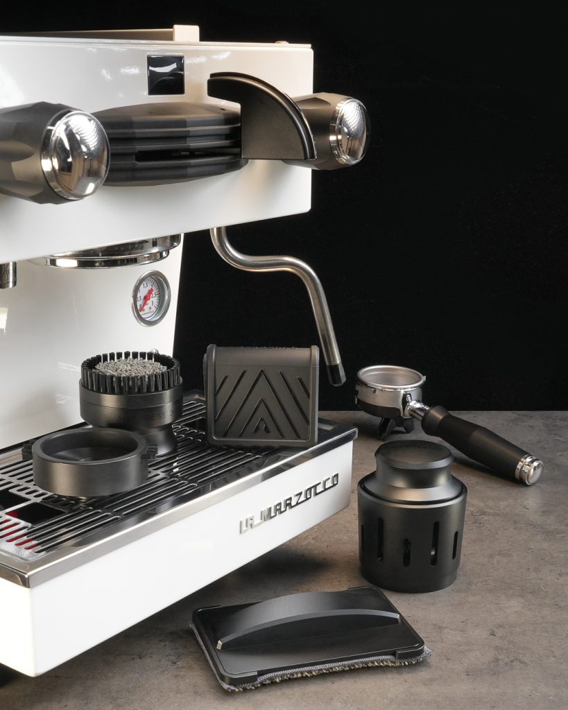 Artpresso cleaning tools arranged on and beside a white La Marzocco espresso machine - group head tool, stone platform, steam wand tool, portafilter cleaning tool and drain tray tool