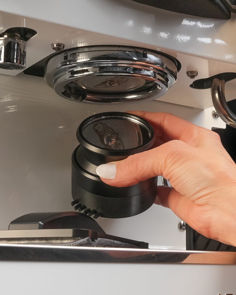 Hands holding the 58mm Group Head Pro cleaning tool below the group head, with the removed shower screen seated inside the cup