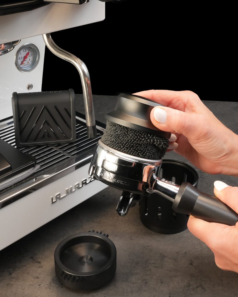 Hands fitting the Portafilter Pro cleaning tool into a portafilter beside a white La Marzocco espresso machine