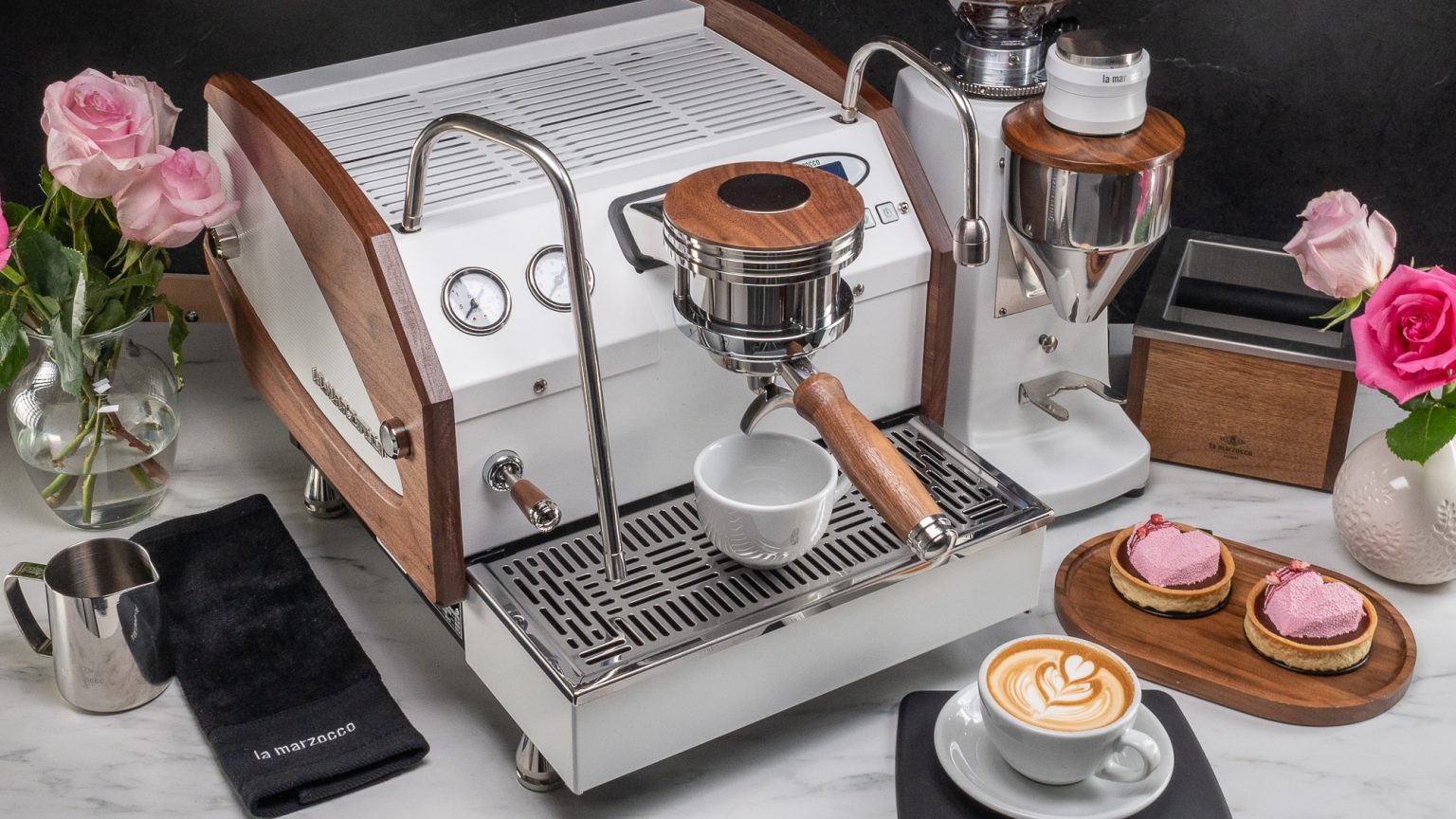 White La Marzocco GS3 with walnut side panels and a walnut portafilter handle on a marble counter, beside a white grinder with a walnut hopper lid and knock box, with roses, a La Marzocco pitcher and towel, two pink tarts and a cappuccino, against a black marble wall