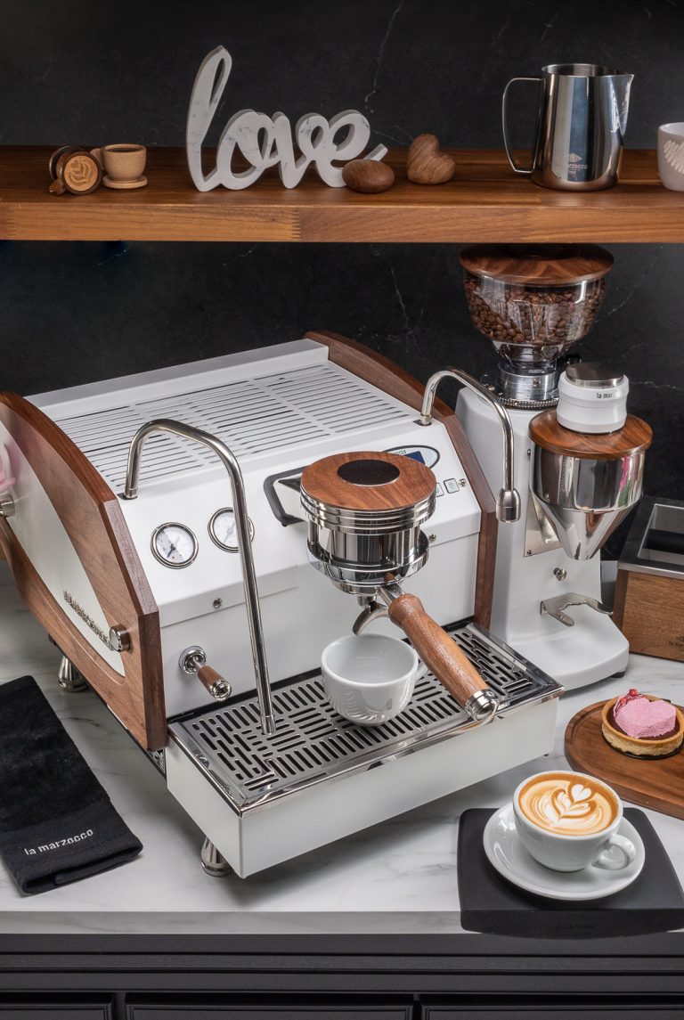 White La Marzocco GS3 with walnut side panels and a walnut portafilter handle on a marble counter, beside a white grinder with a walnut hopper lid, below a walnut shelf holding a La Marzocco pitcher and wooden coffee pieces, with a pink tart and a cappuccino alongside