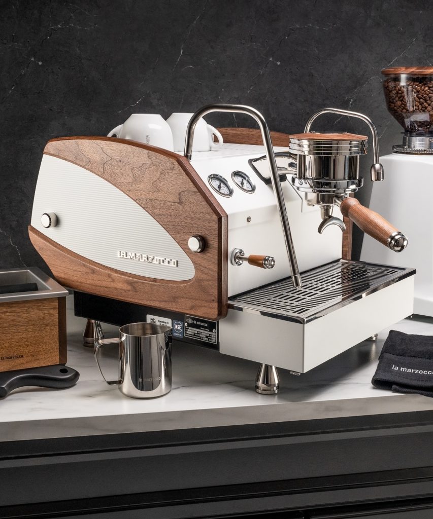 La Marzocco GS3 in white with a figured walnut side panel and walnut portafilter handle, beside a grinder, milk pitchers and cups on a dark counter
