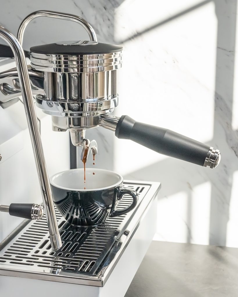 Espresso pouring from a bottomless portafilter with a black handle on a La Marzocco GS3 into a black fluted cup, marble behind