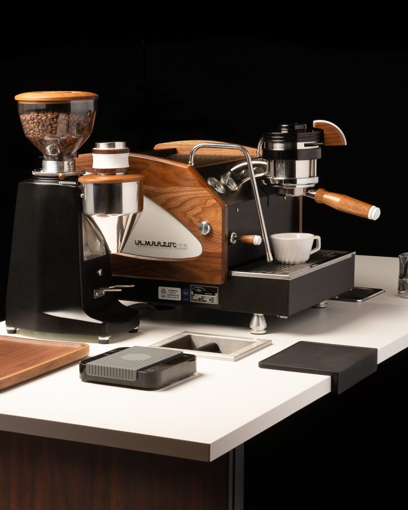 La Marzocco GS3 in black with walnut side panels and a walnut portafilter handle, next to a black grinder with a walnut hopper lid and accessories laid out on a white counter