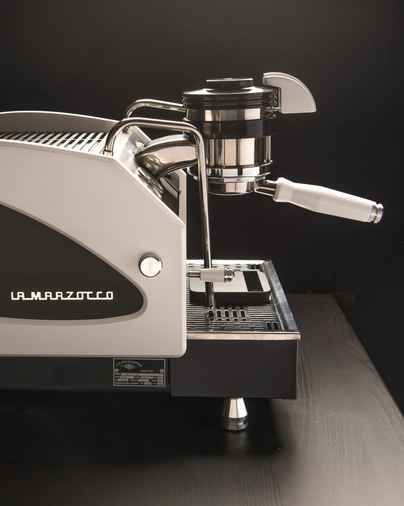 La Marzocco GS3 in white with a black side panel, white portafilter handle and chrome feet, seen from the side on a dark counter