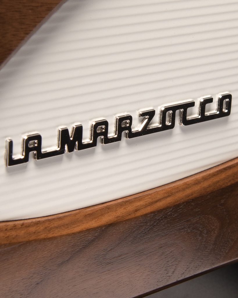 Close detail of the La Marzocco badge on a ribbed white panel with a curved walnut trim running below it