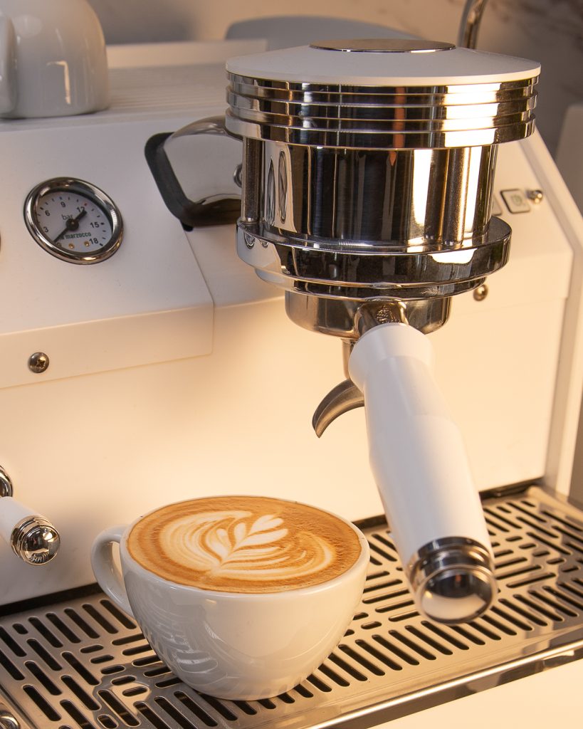 Bottomless portafilter with a white handle locked into the group of a white La Marzocco GS3, a cup of latte art on the drip tray below