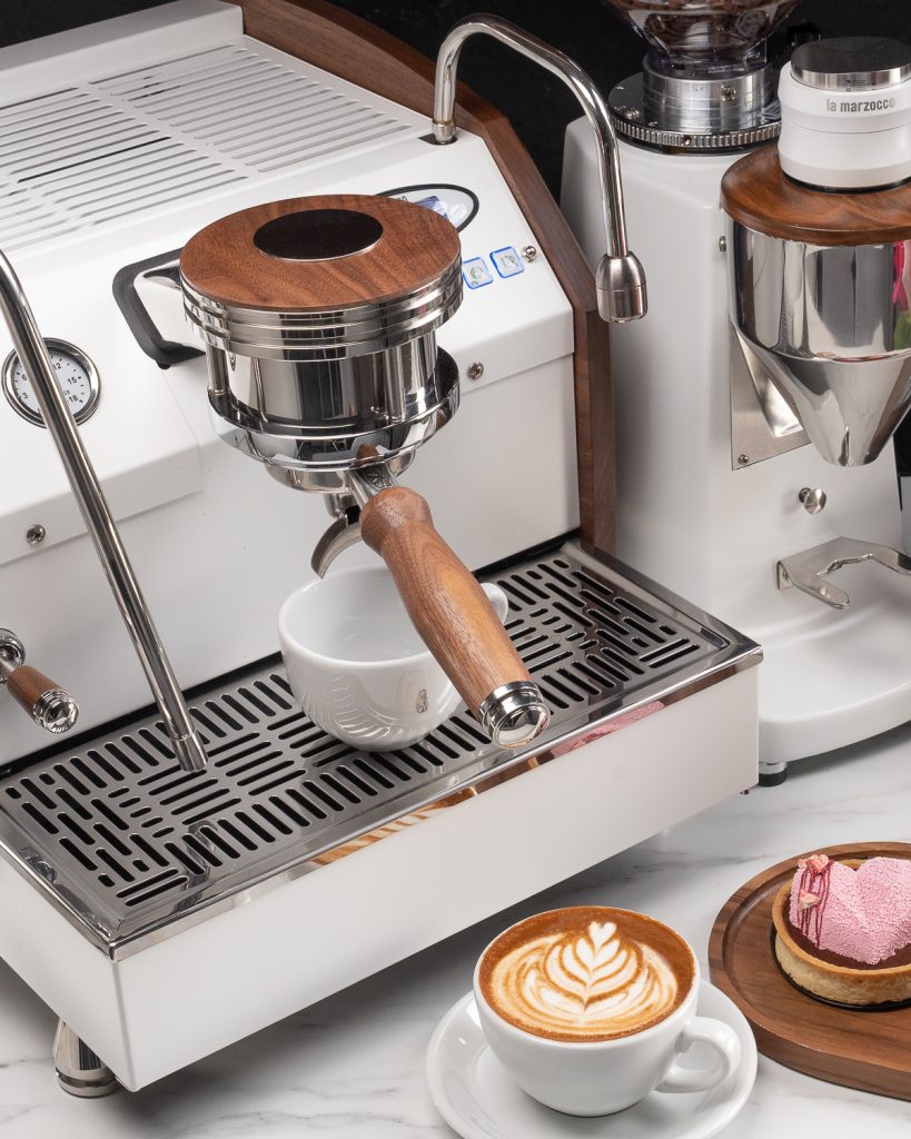 White La Marzocco GS3 with walnut side trim and a walnut portafilter handle, a cup under the group, with a cappuccino and a dessert on a marble counter