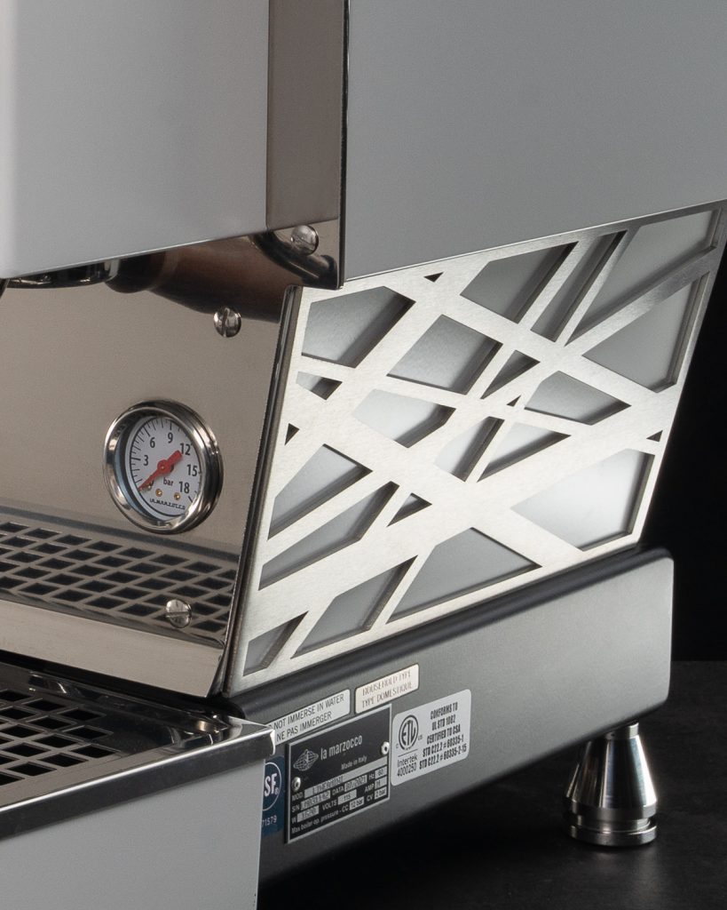 Close detail of a brushed metal cut-lattice side panel standing proud of a La Marzocco Linea Mini, with the pressure gauge and drip tray alongside