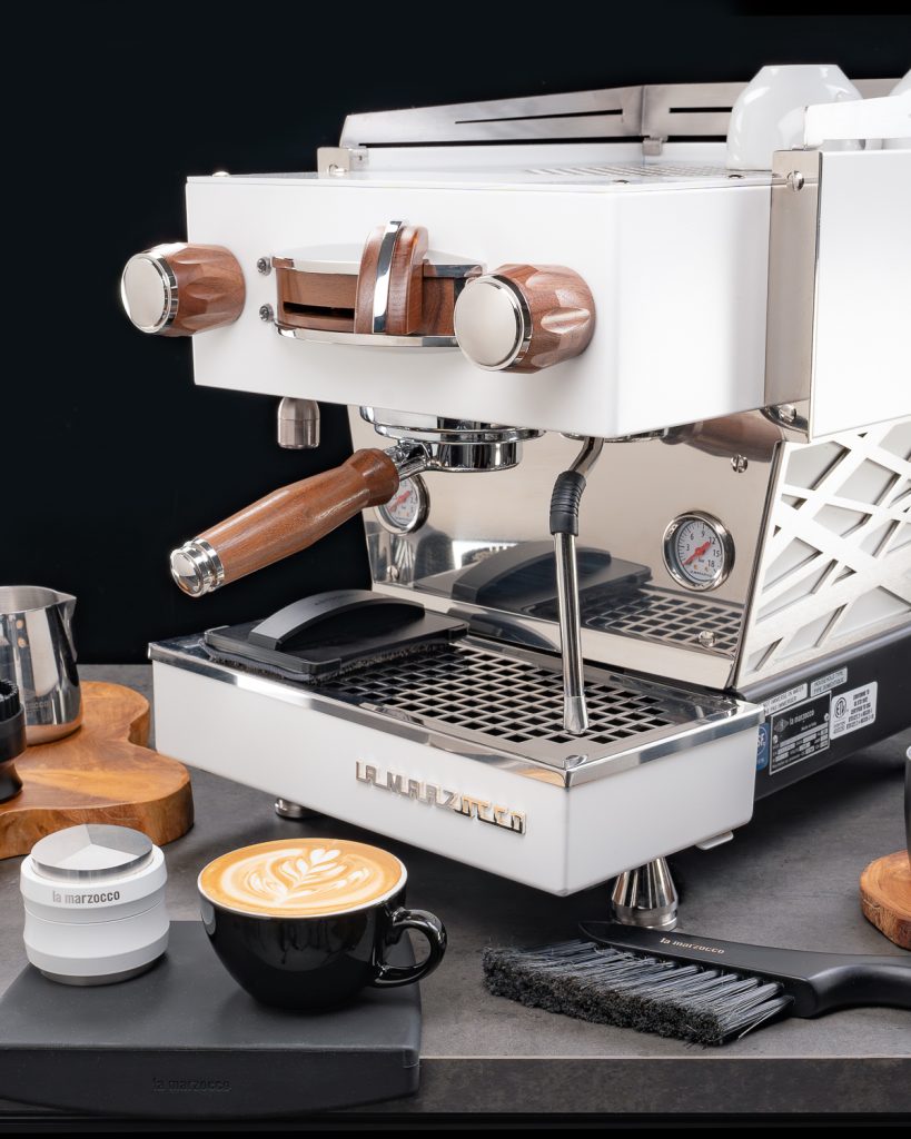 White La Marzocco Linea Mini with walnut steam knob, brew paddle and portafilter handle, on a dark counter beside a cappuccino, milk pitcher and dosing cup
