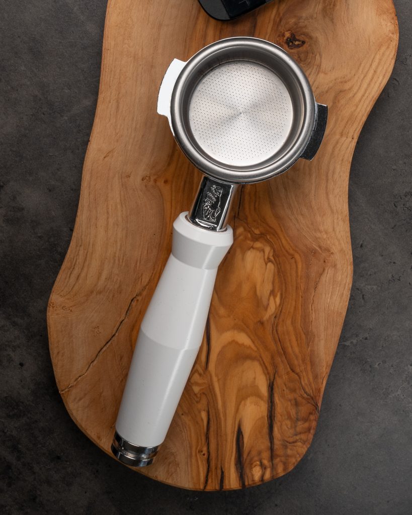 Portafilter with a white handle and a fitted basket, seen from above on an olive wood board