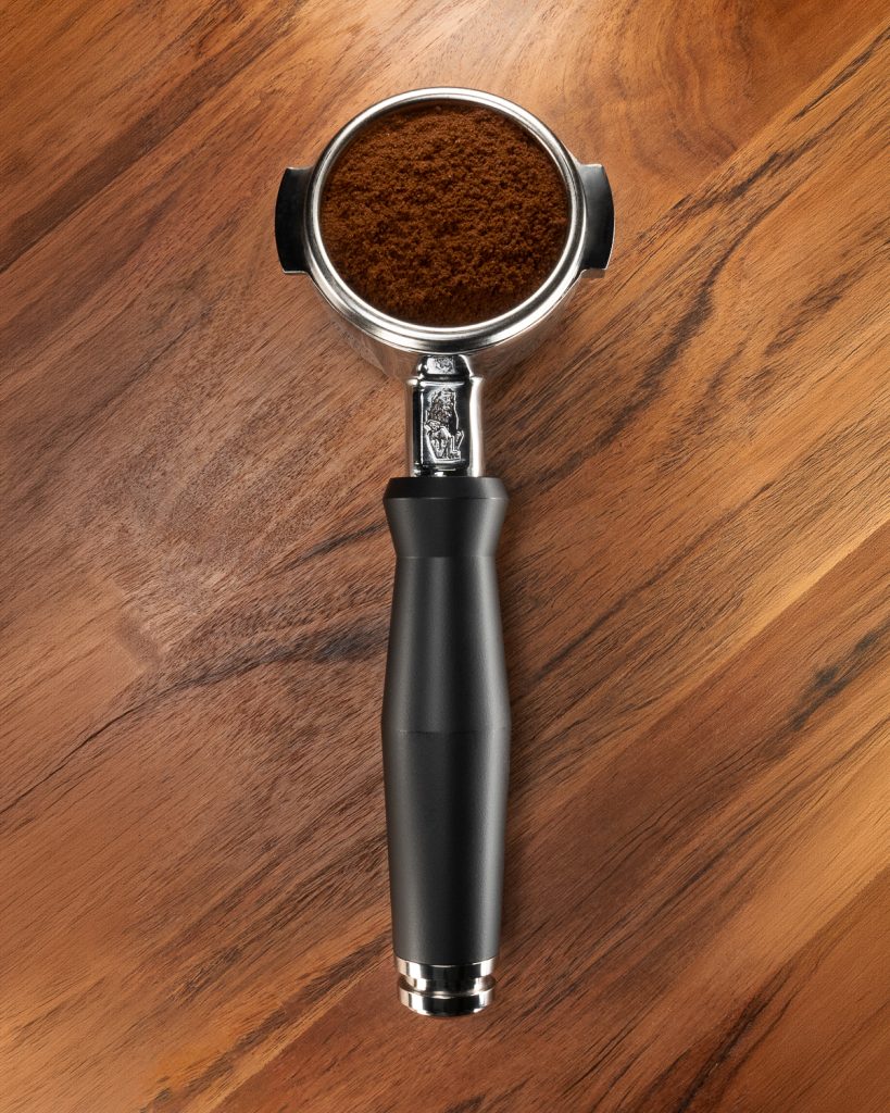 Portafilter with a black handle, its basket filled with ground coffee, seen from above on a wood surface