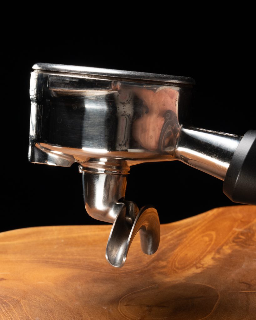 Side view of a chrome portafilter with a black handle and a single spout, against black with warm wood below
