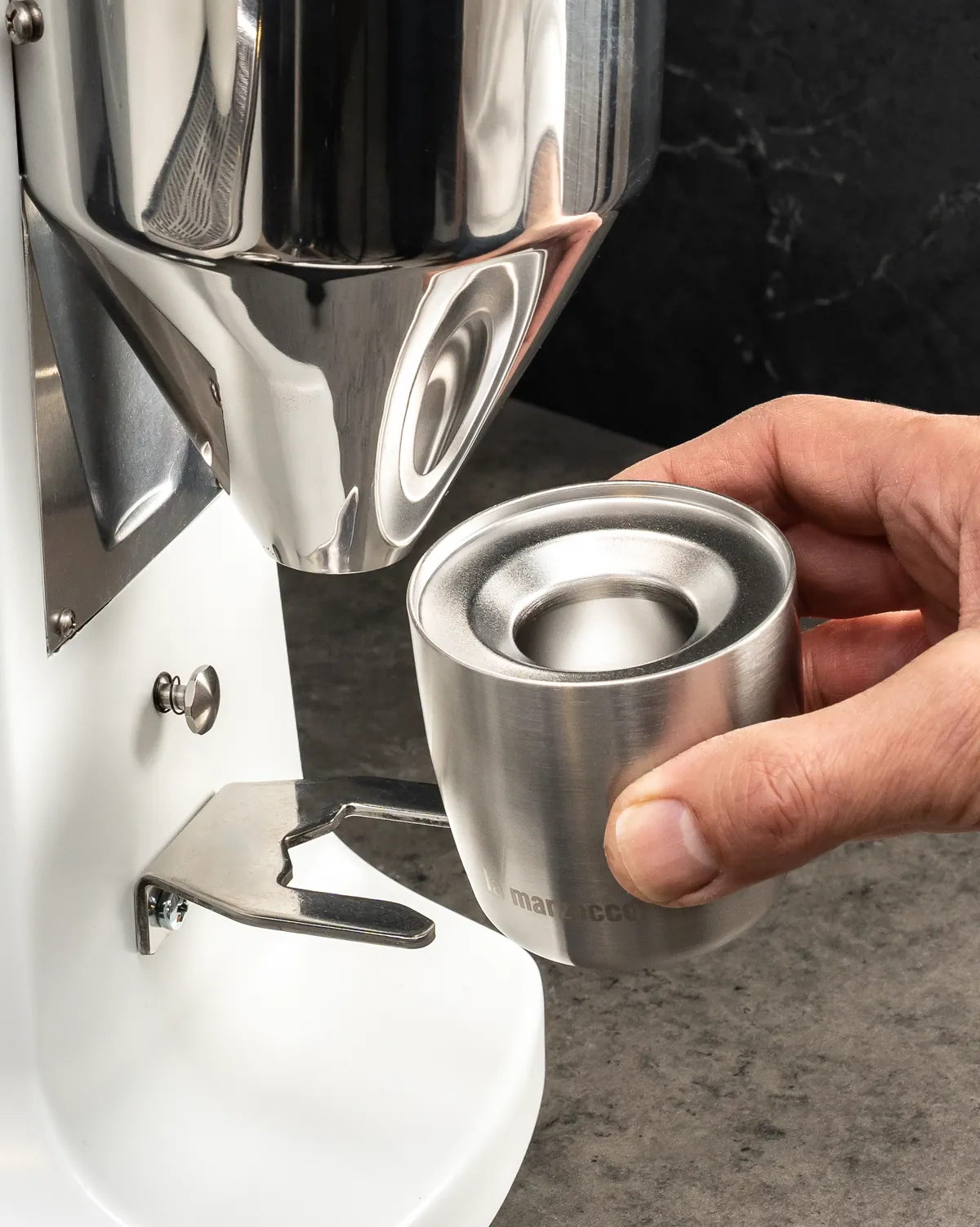 Hand holding a stainless steel dosing cup beneath the spout of a white espresso grinder