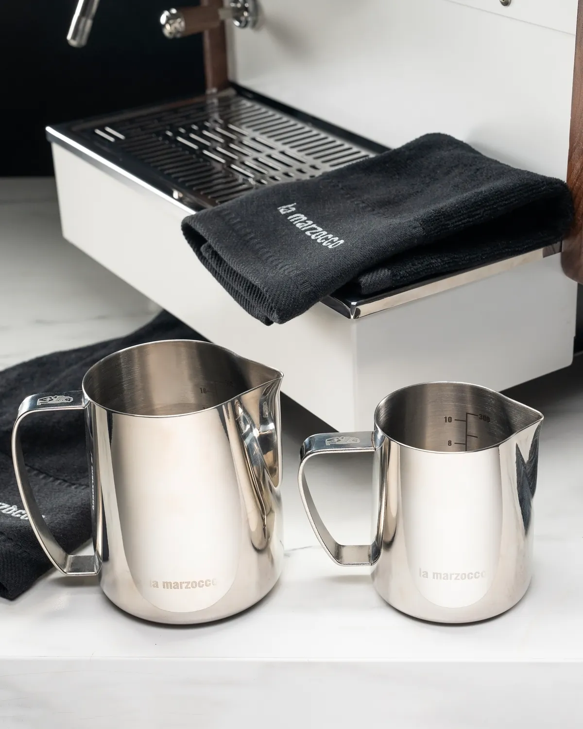 Two stainless steel La Marzocco milk pitchers on a counter beside a black bar towel