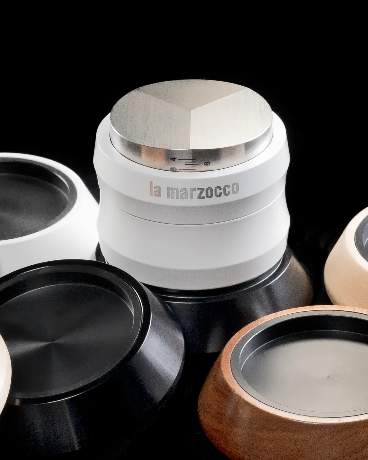 White La Marzocco coffee leveller with a faceted steel top, beside black and wooden lids
