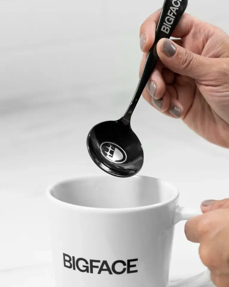 Black BIGFACE cupping spoon held above a white BIGFACE mug