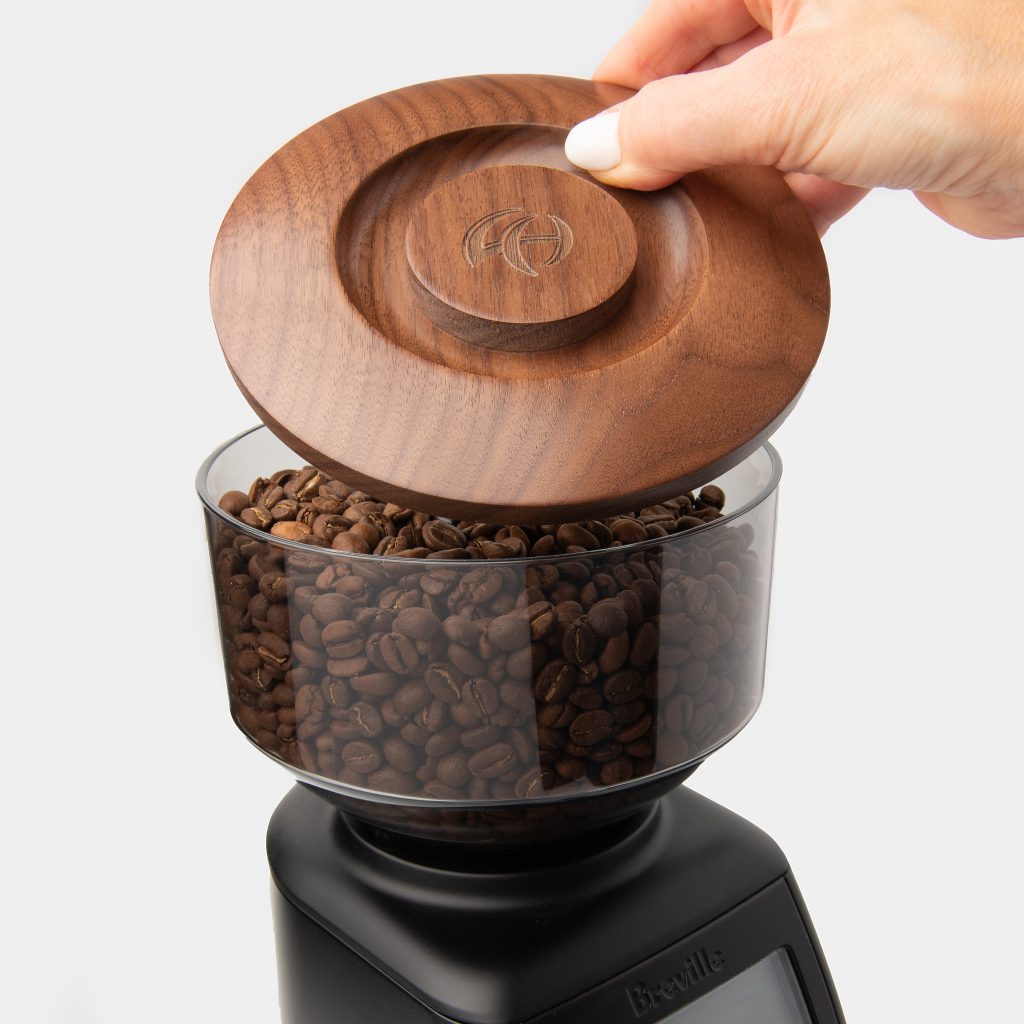 Hand lifting the walnut Breville grinder hopper lid off a hopper filled with roasted coffee beans