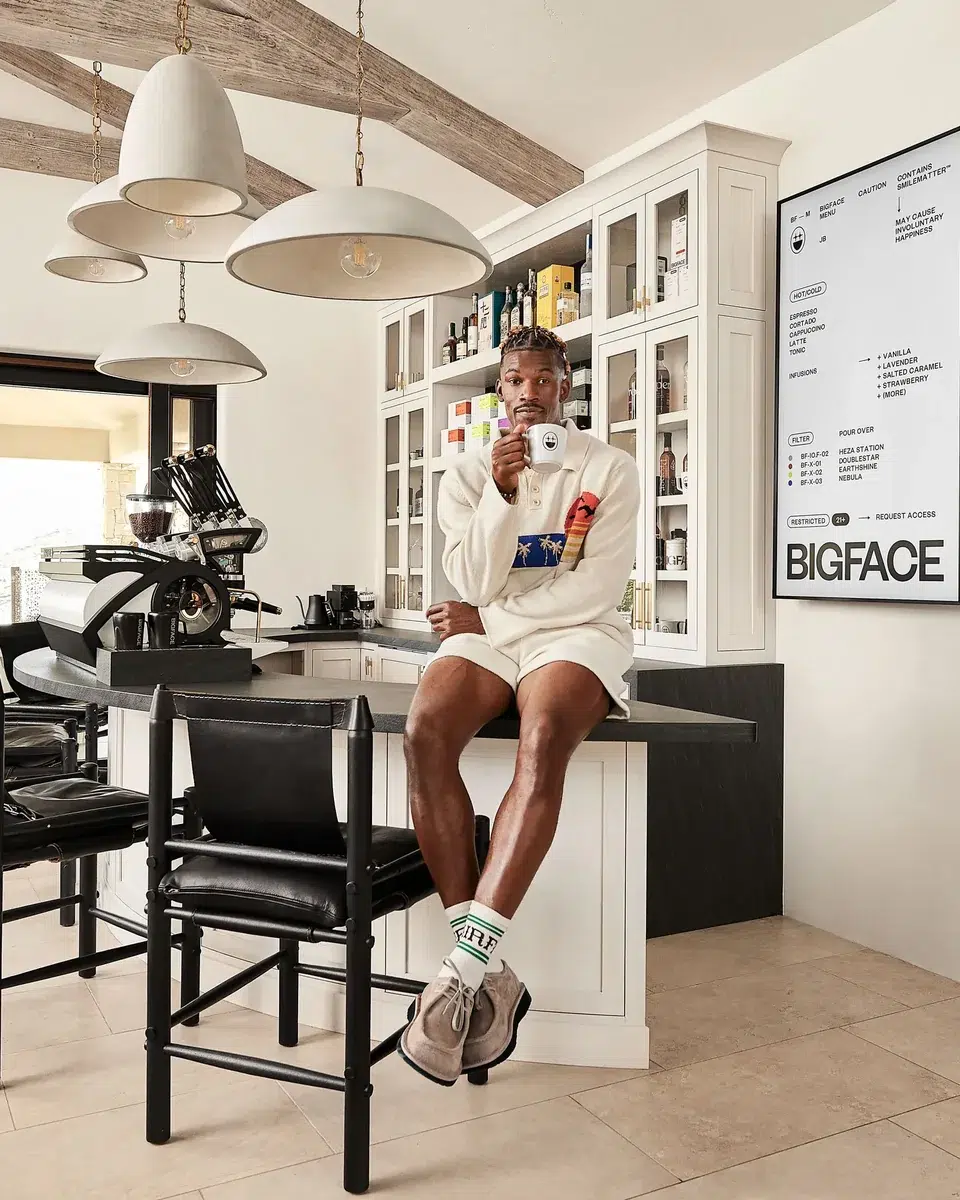 Jimmy Butler seated on his kitchen counter at home holding a BIGFACE cup, with the commissioned La Marzocco Leva behind him