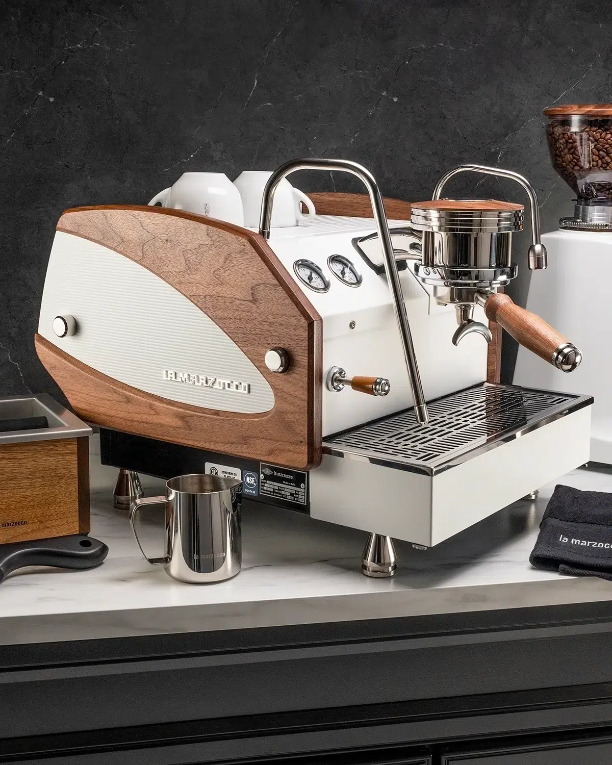 White and walnut La Marzocco espresso machine with a wooden portafilter handle, beside a grinder and walnut knock box