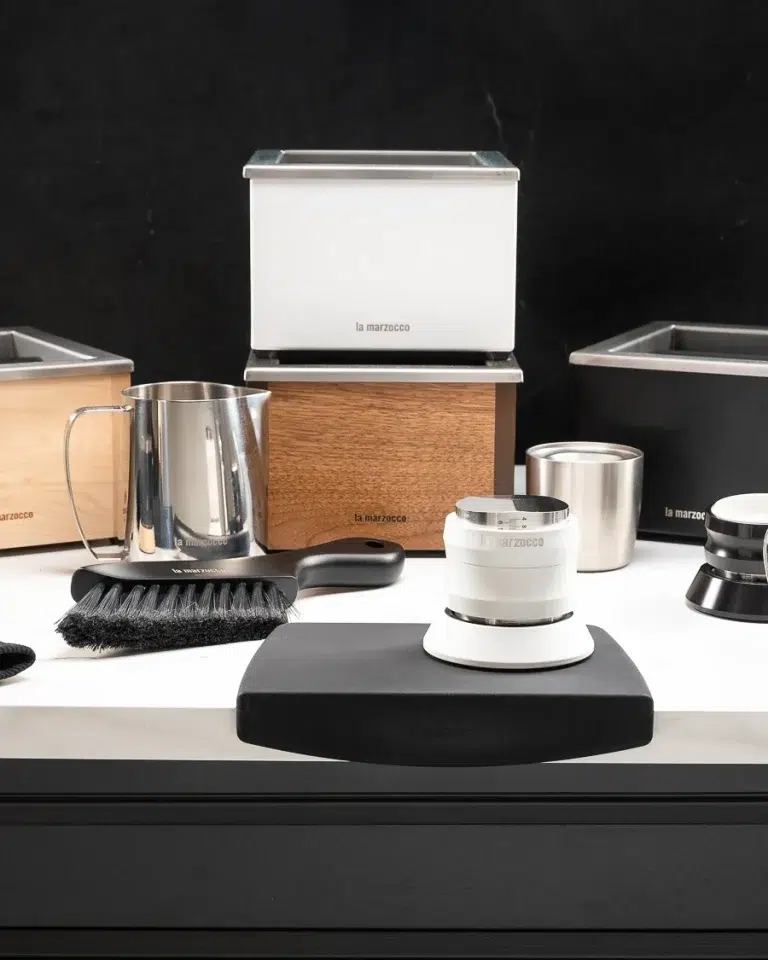 White La Marzocco knock box stacked on a walnut knock box, with a maple box, milk pitcher, barista brush and white distribution tool on a black tamping mat