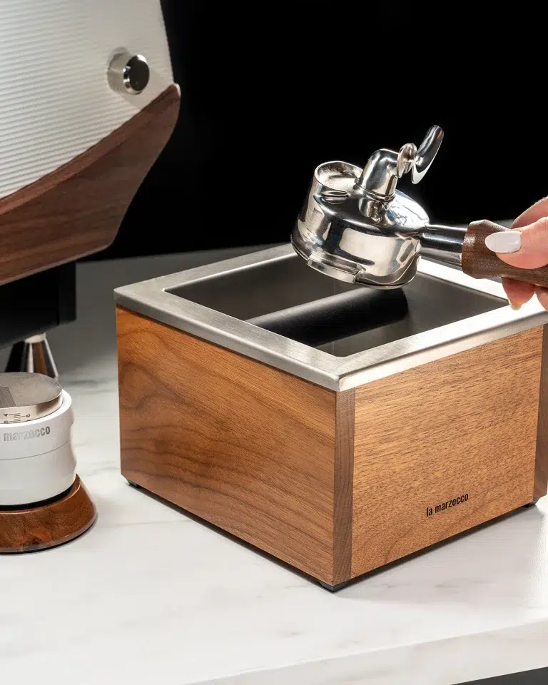 Barista knocking a portafilter out into a walnut La Marzocco knock box beside a white espresso machine