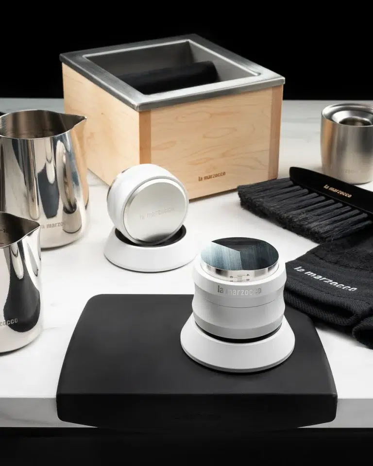 Maple La Marzocco knock box with two white distribution tools, milk pitchers, a barista brush and stainless dosing cup on a black tamping mat