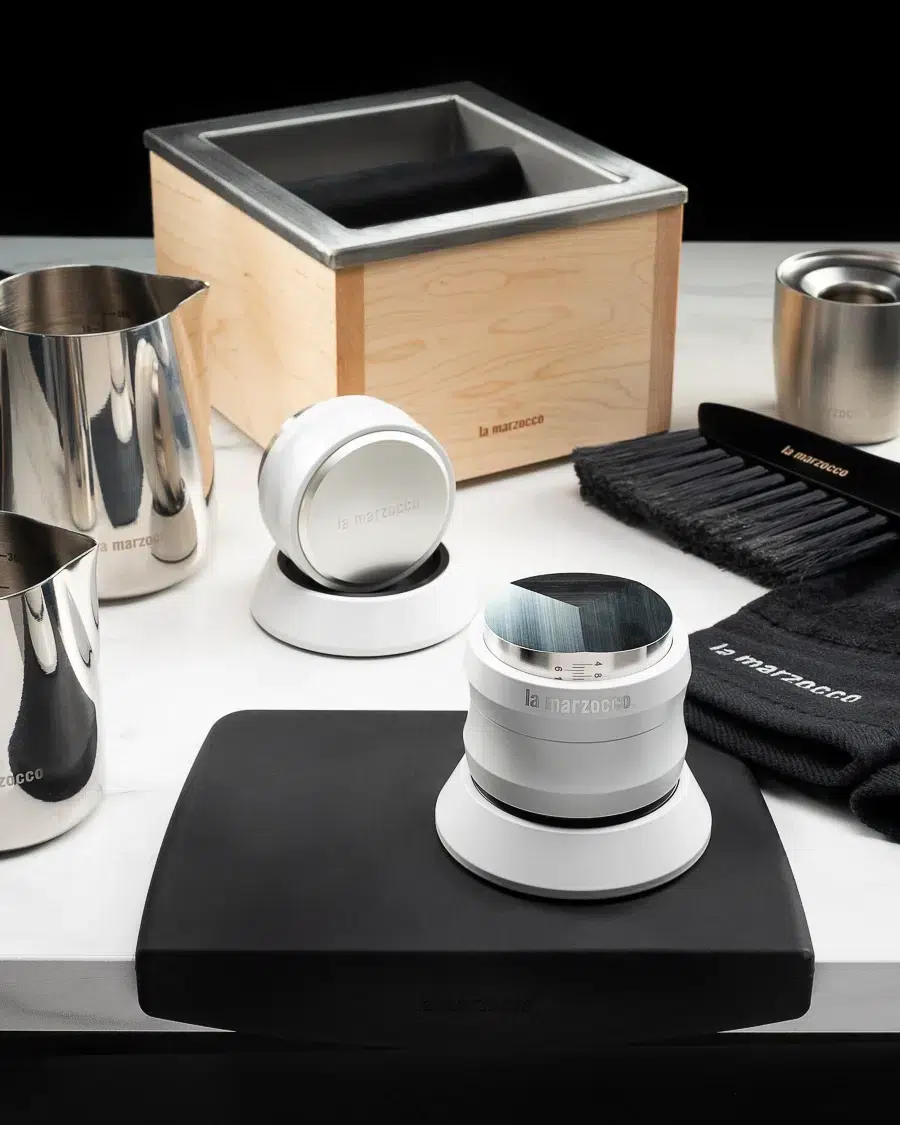 Maple La Marzocco knock box with two white distribution tools, milk pitchers, a barista brush and stainless dosing cup on a black tamping mat
