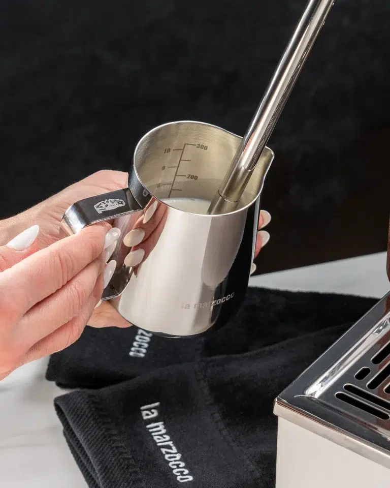Barista steaming milk in a stainless La Marzocco pitcher, with a black branded towel on the counter