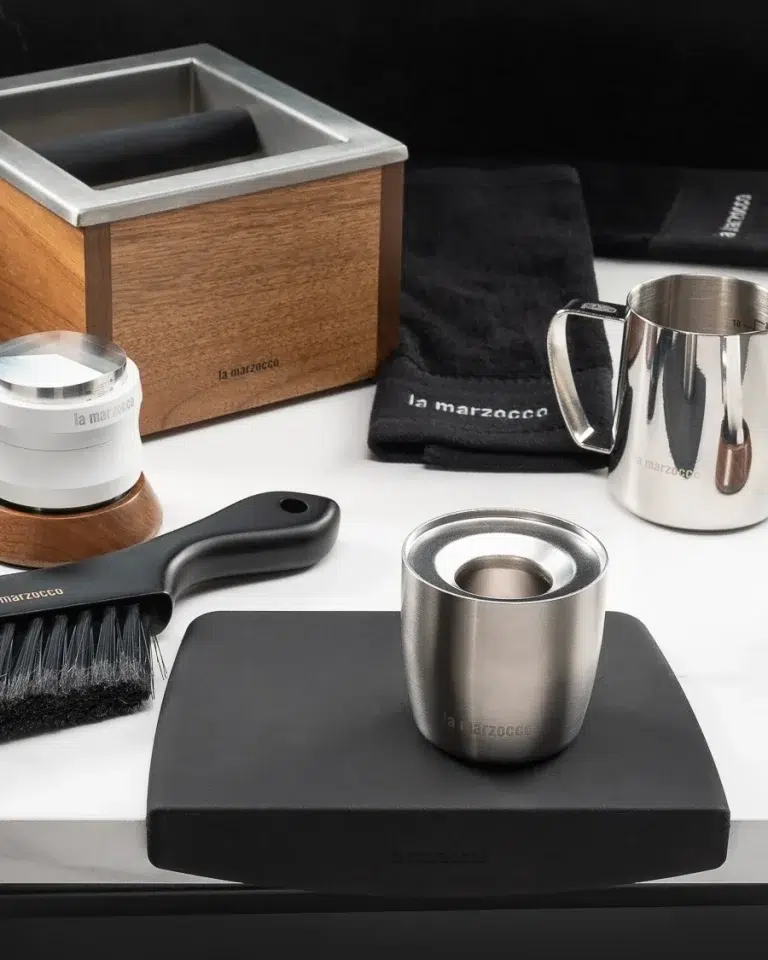 Walnut La Marzocco knock box with a white distribution tool, barista brush, stainless dosing cup and milk pitcher on a black tamping mat