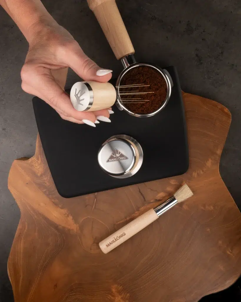 Hand using the maple Mahlkönig distribution tool on a dosed portafilter, with the brush and dosing cup on a live edge walnut board