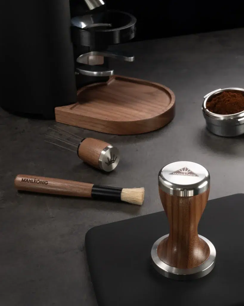 Walnut Mahlkönig tamper, brush, distribution tool and dosing tray on a dark counter beside the grinder