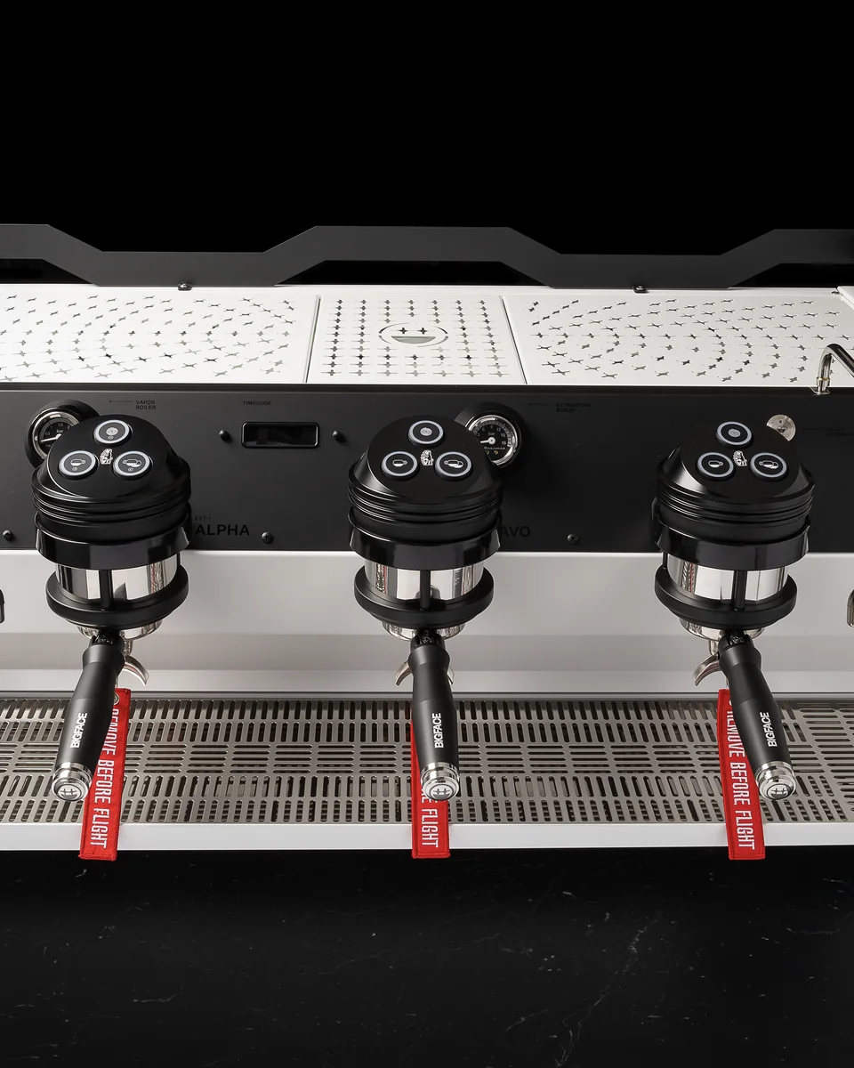 Three group heads of the custom BIGFACE La Marzocco Strada with BIGFACE portafilters and red REMOVE BEFORE FLIGHT tags, beneath the star-perforated cup tray